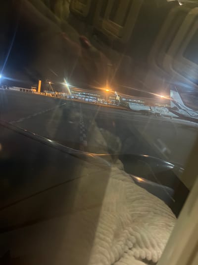 The image, taken from an airplane window at night, shows an airport tarmac with several aircraft parked. The terminal building is visible in the background, illuminated by lights. One of the planes has "Enugu Air" written on its side, suggesting it is part of the Enugu State airport operations. The foreground shows a portion of the airplane wing and the interior of the aircraft cabin, including a blanket. The scene is dark with artificial lighting from the airport and aircraft. Based on the presence of "Enugu Air" and the visual context of an airport, the location is likely Enugu State Airport, Nigeria, not Gwaska.
