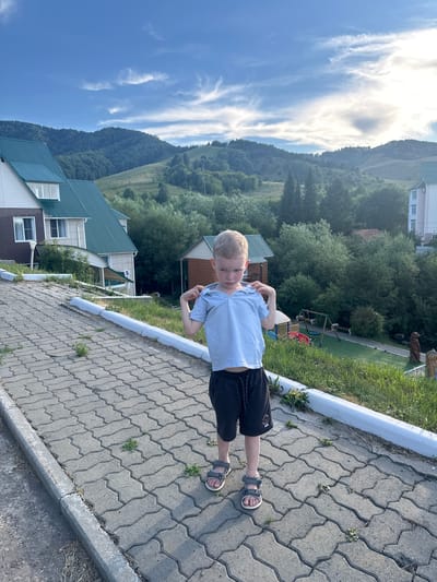 A young boy stands on a paved pathway in what appears to be a resort area. He is wearing a light blue polo shirt and dark shorts, with grey sandals on his feet. The boy is looking down and holding the collar of his shirt with both hands. The pathway is made of interlocking grey pavers and slopes gently uphill. To the left of the pathway, there are several buildings with green roofs. In the background, lush green hills and trees are visible, with a cloudy sky overhead. To the right of the pathway, there is a playground with swings and a slide, and further back, a large building. There are also some decorative bear statues visible on the right side of the image. The overall impression is a scenic, outdoor location likely in a mountainous region, consistent with the provided location information of Danilovka (Altai), Russia, known for its natural beauty and recreational facilities.