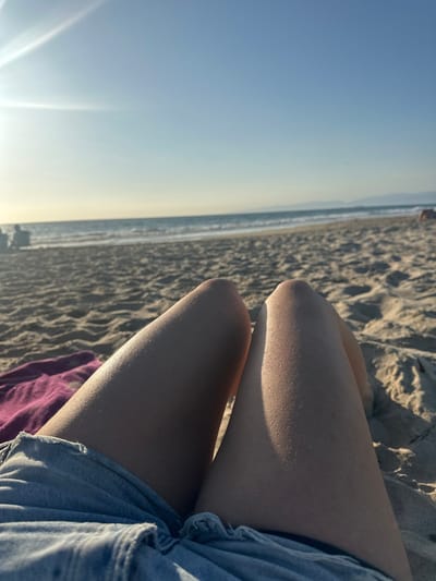 The image depicts a first-person view of a sandy beach in Manhattan Beach, United States. In the foreground, the bare upper legs of an individual, partially clad in light blue denim shorts, are visible, with prominent sunlit highlights on the right leg. To the left of the legs, a crumpled dark reddish-purple textile object rests on the light-colored sand. The mid-ground transitions from the sandy beach to the Pacific Ocean, where small waves are breaking. Multiple indistinct human figures are discernible on the beach in the far left background near the waterline. The background features a clear, light blue sky with a bright sun flare in the upper left quadrant, indicating a low sun angle. Faint dark landforms are visible on the distant horizon to the right.