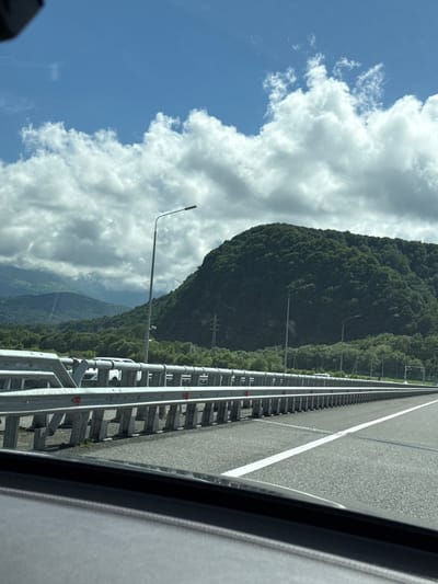 A view from a moving vehicle on a highway in Alagir, Russia, showcases a picturesque landscape dominated by lush green mountains under a partly cloudy sky. The highway features a metal guardrail system on the left, with a clear white lane marker dividing the asphalt road. In the distance, dense forests cover the rolling hills, leading up to a prominent, rounded mountain peak. Streetlights are visible along the roadside, suggesting an organized infrastructure. The overall scene is one of natural beauty combined with modern transportation, typical of mountainous regions.