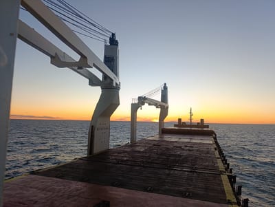 The photograph captures a wide-angle view of the deck of a cargo ship at sea during sunset. The sky is a gradient of clear blue at the top, transitioning to pale yellow and then a vibrant orange at the horizon. The sun is a bright, small disc just above the horizon line. The sea is a deep blue, with small waves rippling across its surface, reflecting the colors of the sunset.

Dominating the foreground and mid-ground are the ship's large, white cranes. The perspective is from the deck, looking towards the stern and out towards the open water. The deck itself is a dark, weathered surface, possibly made of wood or a similar material, with visible seams and fasteners. Yellow markings are present along the edge of the deck. On the left side of the frame, a large white support structure for a crane is visible, with the text "NO SMOKING" printed vertically on its side. Cables from the cranes extend upwards and across the sky. In the distance, the silhouette of another crane structure and parts of the ship's superstructure are visible. The scene conveys a sense of open space and the vastness of the ocean. The location in Larsmo, Finland, is not directly evident from the visual elements of the image, as it depicts a maritime scene at sea, which could be anywhere.