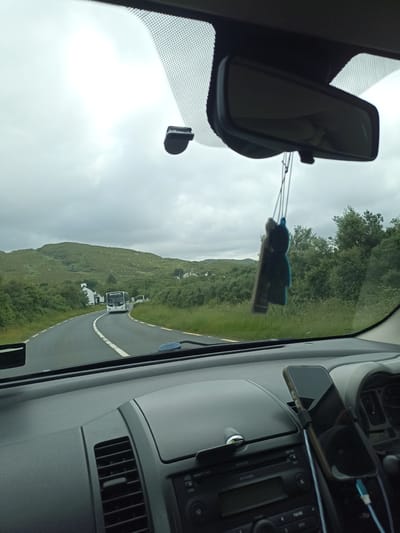 The image captures a view from inside a car driving on a winding road in Crolly, Ireland. The scene is dominated by lush green rolling hills covered in vegetation, typical of the Irish landscape. A cloudy sky suggests overcast weather.
In the distance, a white bus is approaching the car on the road. The road itself is asphalt with white and yellow lines indicating lanes. There are scattered houses and buildings visible amidst the greenery, hinting at a rural or semi-rural setting. Dense green trees line the sides of the road, creating a natural tunnel effect in some areas.
Inside the car, the dashboard is visible, featuring a car stereo and an air vent. A black car accessory, possibly an air freshener shaped like a figure, hangs from the rearview mirror. A smartphone is mounted on the dashboard, its screen reflecting the car's interior. The time of day appears to be daytime, given the ambient light, but the overcast sky contributes to a somewhat muted light. The overall impression is a scenic drive through the Irish countryside under a cloudy sky.