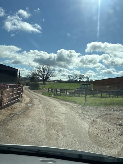 The image depicts a rural scene in Pulverbatch, United Kingdom. A dirt road runs through the frame, flanked by green fields and trees. A tractor is visible in the middle distance. The sky is blue with scattered white clouds. The image is likely taken from inside a vehicle, as part of the vehicle's interior is visible in the foreground.