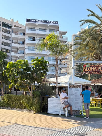 This image depicts a sunny outdoor scene at a beach club. The foreground features lush green foliage and palm trees, with the buildings of a resort complex visible in the background. Two men are present, one wearing a blue t-shirt and shorts, and the other in a white t-shirt and shorts. They appear to be interacting with or observing a menu board. The menu boards advertise "ALOHA PLAYA BEACH CLUB" and "APARTAMENTOS ODYSEA." The setting suggests a warm, daytime atmosphere, likely in a coastal tourist area. The overall impression is one of a casual, relaxed vacation environment.