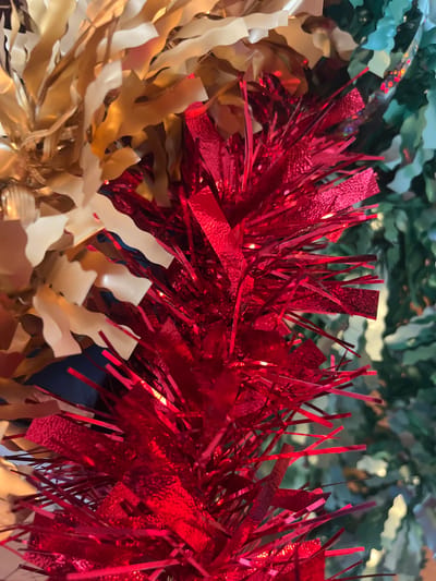 This close-up image depicts a dense arrangement of festive decorations, located in the city of Callbach, Germany. The central and lower-right areas are dominated by bright red tinsel, composed of narrow, reflective, metallic strips. Interspersed within the red tinsel are small, indistinct points of light. To the upper left, a section of gold-colored garland is visible, featuring wider, wavy metallic strips with a matte to semi-gloss finish. On the far right, a blurred green element, consistent with artificial evergreen foliage or green tinsel, provides a background contrast. No individuals, actions, or specific structural elements of a room are discernible; the focus is solely on the interwoven decorative materials.