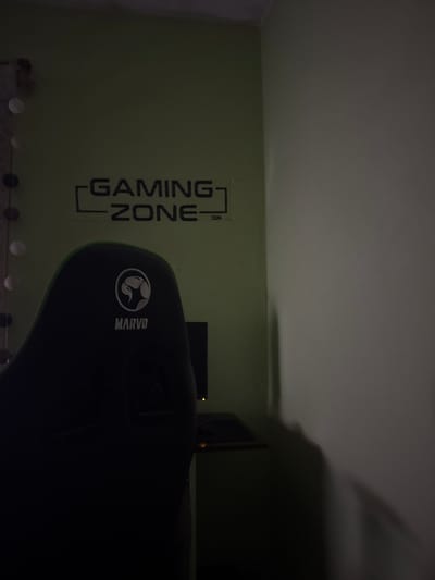 The image depicts a room with a "GAMING ZONE" wall decal. A black gaming chair with "MARVO" branding is positioned in the foreground. A monitor and what appears to be a desk are visible in the background. The location is not discernible from the image alone and therefore cannot be said to be Bratya Daskalovi, Bulgaria, from the image alone. The lighting is dim, with a shadow cast in the upper right corner.