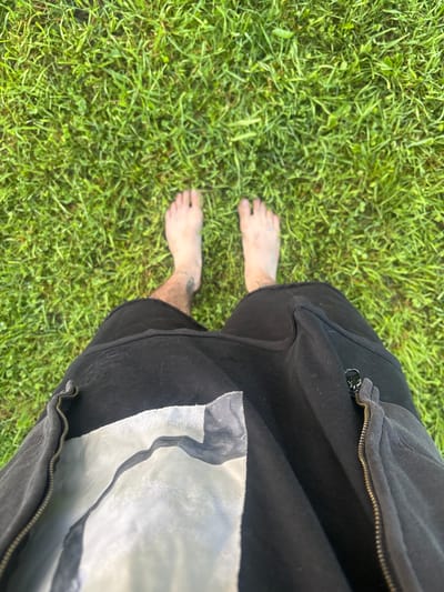 In the city of Rybaki, Russia, a person stands barefoot on a patch of green grass. The perspective is from above, looking down at their feet and the lower part of their legs. They are wearing black shorts, one of which has a zipper and a light-colored graphic design visible. The grass appears lush and well-maintained.
