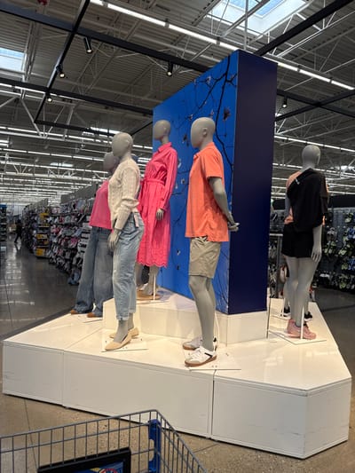 This image, taken in Worcester, United States, displays a retail clothing arrangement featuring five mannequins. The scene is set indoors, likely within a large department or chain store, indicated by the high industrial-style ceiling, exposed structural beams, and track lighting. The store appears well-lit, possibly by both artificial light and natural light filtering through skylights.

The mannequins are positioned on multi-tiered white display platforms against a tall, dark blue panel featuring a light-colored silhouette of tree branches and small birds, suggesting an outdoor or natural theme for the display.

From top to bottom, the mannequins are dressed in various casual outfits:
1.  A mannequin wears a black short-sleeved top, black shorts, and pink sneakers.
2.  A mannequin sports an orange short-sleeved shirt, beige shorts, and white sneakers with brown soles.
3.  A mannequin is dressed in a vibrant pink eyelet-embroidered dress with a cinched waist, paired with light tan flat shoes.
4.  A mannequin wears a light beige long-sleeved knit sweater, blue jeans, and tan slip-on flat shoes.
5.  A fifth mannequin is partially visible behind the fourth, wearing a pink t-shirt and blue jeans.

In the background, out of focus, are numerous clothing racks showcasing merchandise, further confirming the retail store environment. To the right, a metal shelving unit or cart is partially visible. No discernible text is visible on any of the clothing or display elements. The overall impression is a static, well-organized clothing presentation aimed at shoppers.