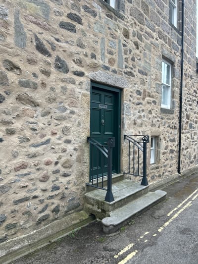 A slightly tilted, medium shot shows the exterior of a building in Aberdeen City, United Kingdom, constructed of irregularly shaped, gray, stone bricks set in mortar. The building has three windows, all with white frames and small panes, visible on the upper portion of the frame. Below the windows is a dark green, paneled door with a silver door knocker. The door is accessed by a small concrete step and flanked by black metal railings on either side. A narrow sidewalk runs along the base of the building, separated from the road by a concrete curb and a double yellow line painted on the asphalt. A black pipe runs horizontally along the upper portion of the building’s facade. Number 25 is partially visible on the wall just above the door.