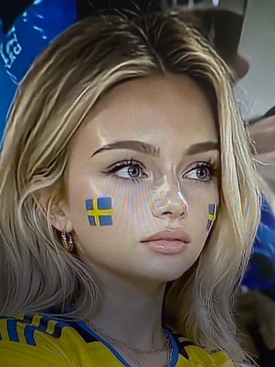 This media file is a close-up, vertically oriented photograph of a young blonde woman's face. She appears to be a spectator at an event, as evidenced by the Swedish flag painted on her cheek and the blue and yellow sports jersey she is wearing. Her hair is styled long and is a light blonde color, with some strands falling across her face. She is wearing makeup, including eyeliner and mascara. The woman's expression is neutral. The background is blurry, but appears to be an indoor setting, possibly a stadium or arena, with hints of blue and yellow patterns. There are no discernible time of day or weather cues. The location context is Chorzów, Poland. There is no visible text in the image.