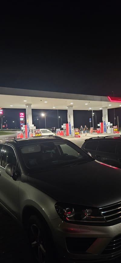 The image depicts a night scene at a gas station in Sobieszczany-Kolonia, Poland. The foreground is dominated by the front portion of a light-colored SUV, its headlights reflecting red light. To the right of this SUV, a dark-colored car is visible. In the background, the gas station canopy is illuminated, with several gas pumps visible. Two men are seen standing near a gas pump, appearing to interact with it. Other vehicles are also present at the station, including a white car parked near a pump. The overall environment is that of a typical gas station at night, with artificial lighting illuminating the structures and vehicles.