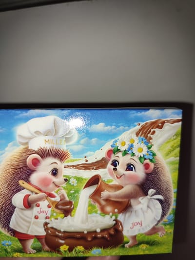 The image depicts an illustration of two anthropomorphic hedgehogs in a grassy meadow under a blue sky with white clouds. The hedgehog on the left, wearing a chef's hat with the word "MILLEN" on it and a chef's uniform with red trim, is holding a wooden spoon and appears to be mixing something in a brown bowl. The name "Jay" is embroidered on its uniform. The hedgehog on the right, adorned with a flower crown, is wearing a white apron with the word "Joy" embroidered on it. It is pouring liquid, likely milk, from a jug into the same brown bowl. The bowl is overflowing with a white frothy substance, and chocolate is depicted as flowing down a slope in the background. The image is presented in a somewhat close-up view. There is no information or visual context in the image that would indicate its location within the city of Voskobiinyky, Ukraine.