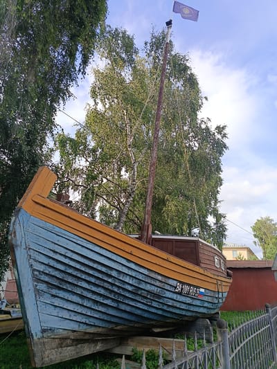 The image shows a wooden boat on display in Shingarskie Isady, Russia. The boat is painted blue and yellow, with visible wear and tear on its surfaces. It is propped up on wooden supports and sits behind a metal fence. A mast with a flag is visible at the front of the boat, and a license plate with the inscription "BA 1001 RUS 35" is attached to the side. Trees and buildings are present in the background, suggesting an outdoor, possibly public, setting. The sky is partly cloudy.