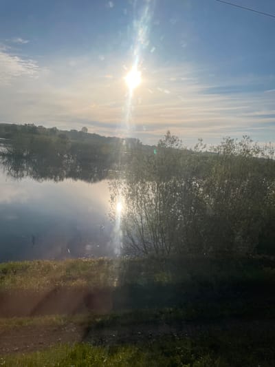 The image shows a bright, sunny scene near a body of water, possibly a lake or river. The sun is high in the sky, causing lens flare. The sky is light blue with long, wispy clouds. The water reflects the sky and sunlight. Along the edge of the water are trees, whose reflections are also visible in the water. On the right side of the image is a grassy area with a dirt path running through it. The details necessary to confirm the location is Talmenka, Russia are not evident within the picture.