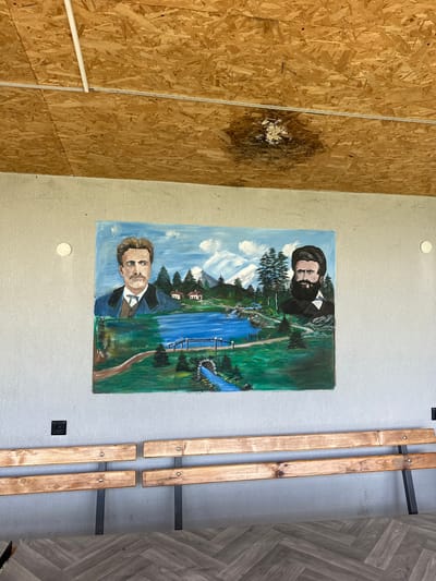 The image shows a wall in what appears to be an interior space in Han Krum, Bulgaria. The wall is painted light gray and has a painting of two men in front of a landscape. The man at the top has a full black beard. The man at the bottom has brown hair. Next to the painted wall is a wall of particle board with what looks like damage. To the right of the painting on the gray wall are two wooden benches with a dark gray metal base. The floor is covered with gray laminate flooring. There are two white circles visible on the painted wall and a black rectangle near the benches.