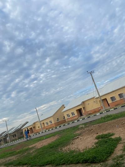 The image depicts a residential street in Dumnga, Nigeria, under a cloudy sky. In the foreground, a patch of unkempt green grass and dry, sandy earth is visible. A paved road runs diagonally across the frame, bordered by a curb with black and white striped markings.
Along the road, a row of yellow houses with white metal roofs is the primary architectural feature. These houses have visible windows and dark doorways. Some of the houses have an orange trim below the eaves. Power lines are strung across the scene, supported by utility poles.
In the middle ground, two individuals, appearing to be male, are walking on the sidewalk in front of the houses. One person is wearing a blue garment, and the other is wearing a dark shirt and shorts. Further down the street, a few more houses with similar architecture are visible, some appearing to be under construction or in an earlier stage of development, with less finished roofs and exteriors. The overall impression is of a developing residential area.
