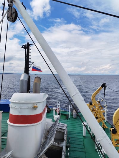 An eye-level, medium shot captures a view from the deck of a vessel on a body of water under a cloudy sky. In the foreground, a large white pipe bisects the frame diagonally from left to right. To the left, a white and red smokestack dominates the mid-ground, with a Russian flag waving from a pole just behind it. A yellow crane is visible to the right of the smokestack. The green deck of the ship extends into the background, meeting the dark blue water. The horizon line is visible in the distance, with a faint outline of land under a sky filled with white and gray clouds. The image appears to be taken in or near Kostyaevka, Russia, given the presence of the Russian flag.