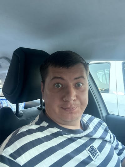 The media file is a selfie taken by a person in a car. The person, likely male, has short, dark hair and is looking at the camera with wide green eyes. They are wearing a blue and white striped t-shirt with a patch on the left chest that has text in an Asian language. The t-shirt also has a small white tag with "AMAZING PLACE" written on it. The person's head is tilted, and they appear to be smiling slightly.
The background shows the interior of a car, including a black car seat and headrest. Through the car window, a white vehicle is visible, suggesting the car is parked or moving slowly. The overall lighting is bright, indicating it is daytime. The scene appears casual and personal, consistent with a selfie taken by someone on the go. There are no clear indications of the weather or specific time of day beyond it being daylight. No other people are visible in the frame. The location context suggests Narbonne, France, but there are no explicit visual cues in the image to confirm this.