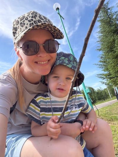 A woman and a young child are in the foreground of this medium shot, with the woman on the left and the child on her lap. The woman is wearing a leopard print baseball cap and sunglasses, and a light-colored t-shirt. She is smiling and looking at the camera. The child is wearing a bucket hat and a striped t-shirt. He is holding a stick and looking away from the camera. Behind them, there is a blue sky with some clouds, a green tree, and a lamppost. The setting appears to be outdoors, possibly a park or a playground, in Kul'dur, Russia. The overall mood of the image is relaxed and happy.