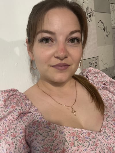 A medium shot photo of a woman looking at the camera. She is wearing a pink floral print dress, a gold chain necklace with a cross pendant, and gold hoop earrings. Her hair is pulled back into a ponytail. The background appears to be a wall with a floral wallpaper. The image does not provide any information about the location in the city of Zubov, Russia.