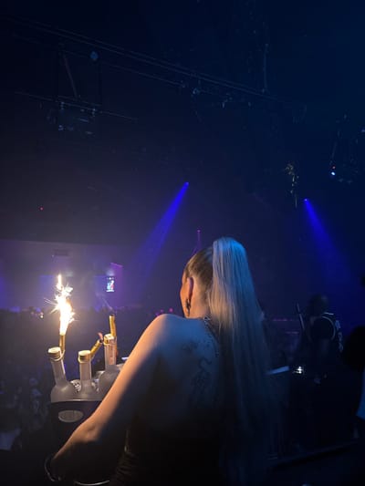 In a dimly lit venue, a woman with long blonde hair and a tattoo on her shoulder is seen from the back. She appears to be at an event where sparklers are being held by multiple bottles in front of her. Blue stage lights illuminate the background, and scaffolding-like structures are visible on the left. A crowd is faintly discernible at the bottom.