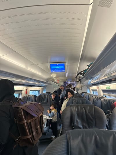 The image displays the interior of a modern train carriage, located in the city of Hattencourt, France, captured from a slightly elevated, forward-facing perspective down the aisle. The carriage ceiling is white and arched, composed of perforated panels, with integrated lighting. An overhead digital display screen in the mid-ground shows blue text "Paris" and "07" on a dark background. Rows of dark grey train seats with headrests are visible on both sides of the central aisle. Windows on the left side reveal blurred outdoor scenery, appearing to be trees.

In the immediate foreground on the left, a person is seen from behind, wearing a dark hooded top and carrying a large, brown leather satchel-style backpack featuring multiple straps and buckles. In the mid-ground on the left, a young child, wearing a light-colored long-sleeve shirt and dark pants, stands in the aisle, looking upwards and to the right. Behind the child, a man with short gray hair is seated in a window seat. Across the aisle, a woman with dark hair and a light-colored jacket is seated in an aisle seat. Further down the carriage, additional passengers are standing and moving in the aisle. On the right side, a blue bicycle is partially visible, secured to an overhead luggage rack. The scene depicts passengers traveling within the train carriage.
