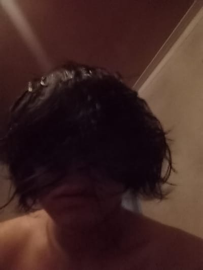 The image captures a close-up, dimly lit scene of a person showering or washing their hair. The primary focus is on the top and back of a head with dark, wet hair. Numerous water droplets are clearly visible clinging to the strands of hair, indicating saturation. A white, translucent stream of water can be seen extending from the upper right toward the hair, suggesting water falling from a showerhead.
Part of the person's bare shoulder and upper back are visible on the left side of the frame, appearing to be light-brown skin, hinting at the person being unclothed, as is typical during showering. The head is tilted downwards, obscuring the face entirely.
The background is dark and somewhat indistinct, consistent with an indoor setting like a bathroom or shower stall. There is a discernible horizontal line towards the bottom right, separating a lighter-colored surface (possibly a shower pan or tub edge) from a darker, reddish-brown wall above it. The lighting is low, creating a soft, intimate ambiance. No specific time of day or location details are explicitly apparent beyond it being an indoor space. There is no visible text in the image.