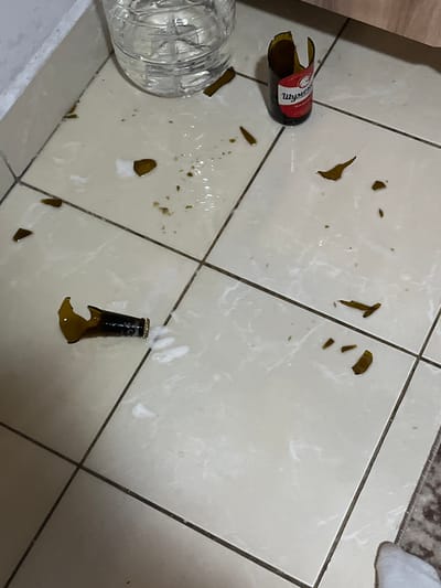 In a domestic setting, likely a kitchen or living area with tiled flooring, a scene of disarray unfolds. The subjects are primarily broken glass from at least two bottles. One bottle, with a red and white label that partially reads "Wyndham," lies on its side, its top half shattered. Numerous shards of amber glass are scattered across the light-colored tiles, some sharp and jagged, others small and fragmented. A clear plastic bottle, possibly containing water, rests on the left side of the frame, partially obscuring some of the broken glass.
In the lower right portion of the image, the base of another broken bottle stands upright, capped and with a small amount of foamy liquid still present, suggesting a recent breakage. The liquid has spilled and pooled in places, creating wet patches on the tiles. The lighting appears to be artificial, indicating the scene might be indoors during either day or night. No people are visible in the frame, and the overall mood is one of accident or neglect. The location is contextually identified as Ivanski, Bulgaria.