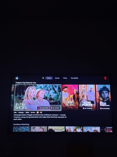 The image shows a Netflix screen on a television, displaying "Today's Top Picks for You". The first suggestion is the film "In Her Shoes". Below it is "Continue Watching". There are several other films and series displayed, including "Desi Bling", "At Home with the Furys" and "Berlin". There is a navigation bar on the left with options such as "Home", "Series", "Films" and "My Netflix". The environment is dark. The image was taken at Aveley, United Kingdom.