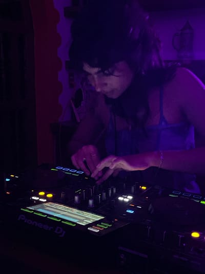 The image depicts an individual, appearing female, operating professional DJ equipment within an indoor, low-light environment in Fortín de las Flores, Mexico. The scene is bathed in a predominant purple ambient light. The individual, with dark hair, wears headphones and a blue-toned spaghetti strap top. Her attention is directed downwards at the equipment, with both hands actively manipulating the controls of a Pioneer DJ controller. Visible jewelry includes a star-charm bracelet on her right wrist and a ring on the ring finger of her left hand. The controller features multiple illuminated buttons, faders, and digital screens displaying track information. The background consists of a reddish wall segment on the left and a darker, less defined area on the right, with faint outlines of what appears to be decorative items on a shelf or wall.