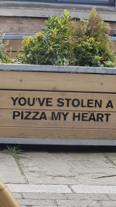 The image shows a vertical view of an outdoor setting. A rectangular wooden planter, filled with green plants, stands adjacent to a paved walkway. The planter has a metal frame. The words "YOU'VE STOLEN A PIZZA MY HEART" are printed in dark capital letters on the wooden side of the planter. The paving stones are light gray and textured. There is no visible evidence of human activity in the frame. The location can be inferred to be Lincoln, United Kingdom from the context.