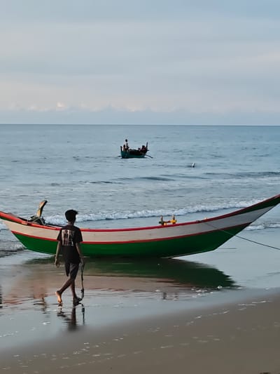 In Peuneulet Baroh, Indonesia, a person walks along a beach toward a colorful fishing boat. The boat, green and white with red trim, rests on the wet sand at the water's edge. The person, wearing a dark t-shirt and shorts, walks away from the viewer, dragging a rope. In the background, another smaller fishing boat with several occupants is visible on the calm sea, with a thin line of clouds on the horizon. The water is a muted blue, reflecting the overcast sky, and gentle waves lap at the shore.