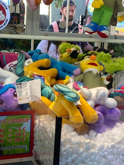 The image shows the interior of a claw machine filled with plush toys. A man is visible through the glass, wearing a black t-shirt and a pink strap across his chest. The claw machine is stocked with various toys, including characters resembling Sesame Street's Bert, Ernie, and Elmo, as well as plush horses from My Little Pony. There is a sign on the left side of the machine with German text that appears to be pricing information. The background suggests an indoor amusement setting or arcade. This image was taken in Baunatal, Germany.