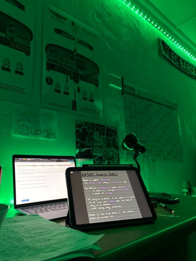 This image depicts a study space located in Port Alfred, South Africa. The room is illuminated by green LED strip lighting mounted along the top of a light-colored, textured wall.

On the wall, from left to right, are two "AIR SCHOOL" posters, one detailing a "Piper Cherokee Archer II / III" cockpit diagram. Below these, a transparent sheet with "Welcome Back Bugs" handwritten in green is affixed. Further right, an "FC BARCELONA" scarf is draped horizontally, and a large "2026" planner poster is visible. Below the planner, another poster titled "Vision Board 2026" is present.

A dark desk occupies the foreground. On the left, a silver laptop is open, displaying an online interface with text and colored indicators. To its right, a dark-cased tablet stands upright, showcasing handwritten notes titled "VDF (VHF Direction Finder)". A stack of papers with writing is situated in front of the laptop. To the right of the tablet, a black pen or stylus lies on the desk. A black flexible desk lamp, emitting a green light, is positioned on the far right of the desk. A clear water bottle or container is visible behind the laptop and tablet.