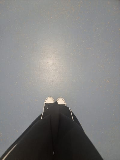 The image displays a downward-angled, first-person perspective of an individual's lower body within an interior setting in Jaunmārupe, Latvia. The subject is positioned upright, with their feet aligned parallel. They wear black trousers featuring a single white stripe along the outer seam of each leg, and black, low-top canvas sneakers with white rubber toe caps, white laces, and white rubber soles. The floor surface is a light grey, mottled with numerous small, irregular yellowish-brown particulates, indicative of a composite material such as speckled linoleum or industrial flooring. A horizontal band of specular reflection from an overhead light source is observable on the floor, located in the upper-mid section of the frame.