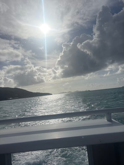 The image presents a daytime marine panorama captured from a moving vessel in the waters off Road Town, British Virgin Islands. In the foreground, the white railing and upper structure of the boat occupy the bottom portion of the frame, with a churning wake visible directly behind it. The expansive blue-green ocean surface is characterized by ripples and sun glint, reflecting the bright sky. A single red conical buoy is discernible in the left-center midground. To the left, a verdant, hilly coastline, consistent with the island of Tortola, recedes into the distance, with some light-colored structures visible on its lower slopes. Further out on the horizon to the right, several low-lying landforms, indicative of other islands, are present, along with a large, dark-hulled vessel. The sky is partially cloudy, featuring both bright white cumulus and darker grey stratocumulus formations. A brilliant, intense sun, exhibiting a prominent lens flare, is positioned in the upper left quadrant, illuminating the clouds and casting a strong reflection on the water. No individuals are visible in the scene.