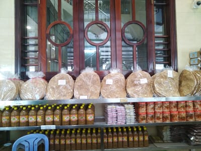 The image shows a shelf stocked with food items, likely in a store in the city of Chu Văn An Ward, Vietnam. On the top shelf, there are stacks of what appear to be large rice paper cakes in plastic packaging, followed by jars of pickled food. The second shelf contains rows of brown liquid in bottles with yellow caps, alternating with stacks of pink and white small cakes. The bottom shelf mirrors the second shelf, with rows of bottles and an assortment of other food packages towards the right. There's a blue plastic chair partially visible in the lower left corner. Behind the shelves is a wall with a wooden window featuring circular designs. A security camera is mounted on the wall near the top of the window.