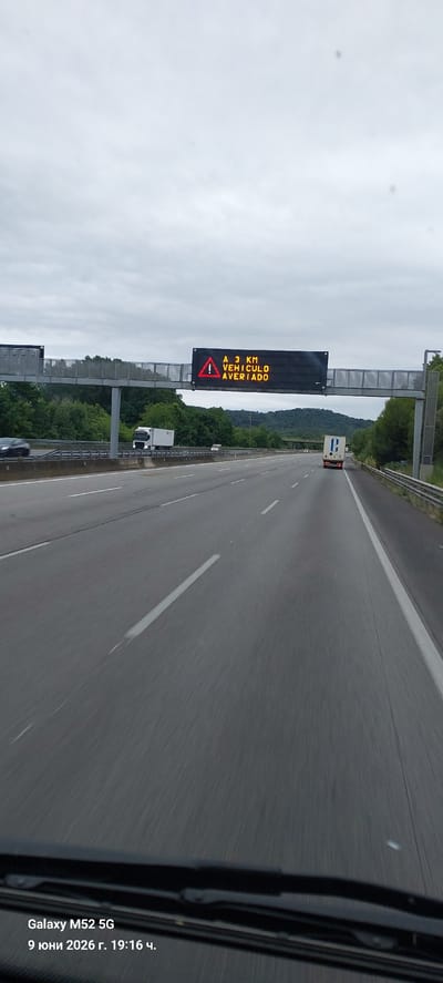 The image captures a view from a vehicle driving on a highway, likely in Sant Julià de Ramis, Spain, given the timestamp. An electronic traffic sign is prominently displayed above the highway, indicating "A 3 KM VEHICULO AVERIADO" with a warning triangle and exclamation mark. This translates to "In 3 km BROKEN DOWN VEHICLE". Several vehicles, including cars and trucks, are visible on the multi-lane highway. The environment appears to be rural, with lush green trees and hills in the background. The sky is overcast, suggesting a cloudy day. The timestamp in the lower-left corner indicates the image was taken on June 9, 2026, at 19:16.