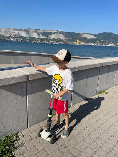 A young boy stands on a paved walkway next to a concrete barrier, holding a small red toy airplane. He is wearing a white t-shirt with a cartoon duck graphic, red shorts with white text, a baseball cap, and gray crocs. He is positioned next to a three-wheeled scooter. The walkway overlooks a body of water with a distant industrial port featuring cranes and buildings. Mountains with a layered quarry landscape are visible in the background under a clear blue sky. The scene is likely the embankment of the city of Novorossiysk, near Sheptal'skii, Russia, given the industrial port and quarry formations.