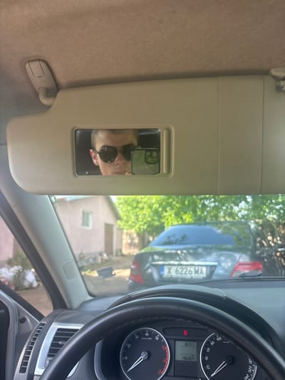 The photograph was taken from inside a car. The dashboard is visible in the lower-right corner of the frame, including a speedometer reading approximately 0 km/h and an odometer reading 196,264. The steering wheel is black leather. Through the windshield, a car is parked behind and to the left, revealing a license plate: "X 6226 MA." The surroundings outside suggest a residential area with trees and houses. In the top-left portion of the frame is the car’s sun visor, and its vanity mirror is reflecting a person in sunglasses and a black shirt. Given the license plate format of "X" followed by four numbers and two letters, the vehicle is likely in Bulgaria, but without further information, it cannot be determined if the location is Oriahovo.
