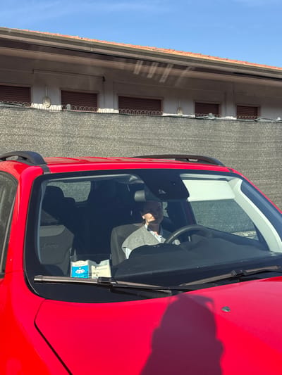 The image captures an outdoor scene in Lipomo, Italy, under a clear blue sky and bright sunlight. The foreground is dominated by the red hood and windshield of a Jeep SUV. Inside the vehicle, a female individual, with dark hair, occupies the driver's seat, looking forward with a faint smile. She wears a light collared shirt beneath a grey sleeveless top; sunlight casts distinct shadow lines across her face. Two white packets or boxes, one with blue details, are visible on the dashboard behind the windshield wipers. In the background, a dark grey mesh fence, topped with a line of razor wire, separates the vehicle from a light-colored building. This building features a terracotta-tiled roof with a gutter system and multiple windows fitted with closed brown roller shutters, interspersed with wall-mounted light fixtures.