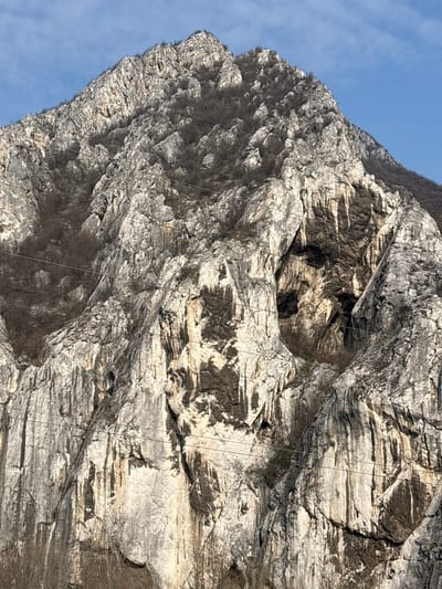 The image displays a large, steep, and rugged mountain face located in Lyutibrod, Bulgaria, under a clear blue sky. The mountain is predominantly composed of light grey rock, exhibiting significant weathering, deep fissures, and vertical dark streaks, likely from mineral deposits or water runoff. Sparse, leafless deciduous trees and bushes cling to the less vertical slopes and crevices, especially visible across the upper half of the mountain.
A prominent natural feature is a large, dark, deeply recessed cavern situated in the central-right area of the rock face, presenting an irregularly ovoid opening. Several smaller, dark cave-like openings are visible nearby. Multiple thin, dark, parallel wires or cables traverse horizontally across the lower and middle sections of the mountain face. No people, animals, or discernible actions are present in the scene. The lighting suggests natural daylight, illuminating the rock formations and the sparse vegetation.