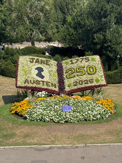 A large, open-book-shaped floral display is prominently featured in the center of the image, located within Parade Gardens in Bath, United Kingdom. The display is constructed from various live plants, forming an open book with two distinct pages. The left page, predominantly light green, spells "JANE AUSTEN" in darker green and reddish-brown foliage, with a central black silhouette of a woman's head in profile. The right page, similarly composed, displays the numbers "1775," "250," and "2025" in green and reddish-brown, signifying a 250-year anniversary. A dark reddish-brown spine connects the two pages. The entire floral book is supported by a dark metal frame.
The book display is situated within a circular flowerbed, primarily filled with white flowers in the foreground and yellow flowers forming a border behind them. A small, rectangular blue sign, partially obscured, is visible among the white flowers, labeled "Bath in Bloom" and indicating "The Jane Austen." The flowerbed is surrounded by a short, dry grassy lawn. A grey paved pathway or road runs horizontally across the bottom of the frame.
In the background, dense green hedges and shrubs create terraced landscaping. Behind these, a thick canopy of mature green trees dominates the upper portion of the scene. Several individuals are visible in the distance, walking or standing on paths. One person, wearing a dark jacket and light trousers, walks towards the left on a path behind the hedges. The scene is illuminated by bright sunlight, casting shadows on the ground.