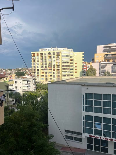 This is a vertical, eye-level shot of an urban landscape in Bashkia Durrës, Albania, captured during the day under a clear, pale blue sky with some wispy clouds. The scene is dominated by a collection of multi-story buildings, varying in color and architectural style, interspersed with lush green trees.
In the foreground, a portion of a light-colored building with a large, grid-like window facade is visible on the right. Adjacent to it, a pale yellow building rises, characterized by rows of balconies and numerous windows. Further back and to the left, more residential buildings are seen, some with cream-colored exteriors and others in lighter hues. The buildings climb up a gentle slope, creating a dense urban environment. The overall impression is one of a well-populated, modern city with a mix of older and newer structures.
On the right-hand building, white text on a red background reads "SHKOLLA 9 VJEÇARE 'A'". This indicates the presence of a school. Electrical wires are visible, running diagonally across the frame, suggesting active infrastructure. The presence of trees and the general brightness of the scene suggest it's a pleasant day, likely late morning or afternoon. No people are clearly visible in the image.