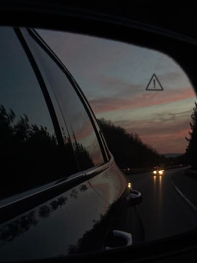 The image captures a view from inside a vehicle at dusk, located on a road near Gorna Vasilitsa, Bulgaria. The left portion of the frame shows the dark, reflective exterior of the vehicle's passenger-side door and window, reflecting the twilight sky and silhouetted trees. The right side is dominated by the passenger-side mirror, displaying a curving asphalt road. In the distance, two sets of bright yellow headlights from oncoming vehicles are prominently visible. Faint yellow-orange light from a closer vehicle or reflector is also present on the right lane. Dark tree lines flank the road, with subtle mountain forms discernible against the horizon. The sky, visible both directly and in reflection, exhibits soft pink and blue hues characteristic of twilight. A prominent triangular warning symbol containing an exclamation mark is superimposed in the upper center of the side mirror's view, appearing as a digital overlay or a reflection from within the vehicle's system. No individuals are explicitly visible.