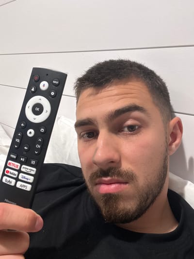 A man is holding a black Hisense TV remote control in his right hand. The remote has buttons for power, input, settings, volume, channel, menu, and quick access to streaming apps such as Netflix, YouTube, Disney+, Prime Video, and Tubi. The man has short, dark hair and a beard. He is wearing a black shirt and lying on a white pillow. The background is a white wall with horizontal lines. The image was likely taken indoors. The location is impossible to determine.
