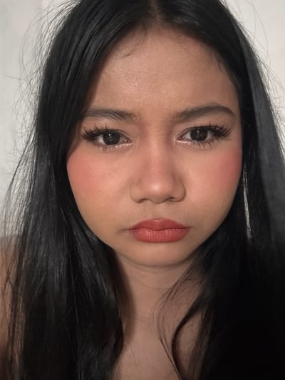 A young woman with black hair and makeup is looking directly at the camera. She is wearing blush on her cheeks and lipstick on her lips. Her hair is long and straight, and it frames her face. The background is a plain white wall. The lighting is soft and diffused, creating a gentle glow on her skin. The image appears to be a selfie, taken at close range, likely in an indoor setting. The woman is the sole subject in the image, and there are no other objects or people visible. The scene does not contain any specific indicators of a North Jakarta, Indonesia location.