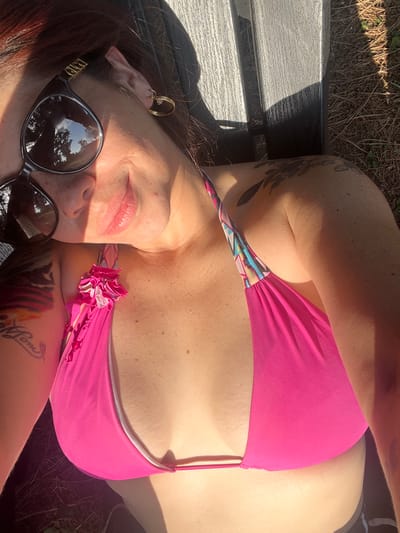 A woman with red hair lies on a bench, sunbathing. She wears a pink bikini top and sunglasses. Tattoos adorn her arms and upper chest. A gold hoop earring is visible in her left ear. Sunlight dapples her skin. Her face is partially in shadow, but her lips are slightly parted in a relaxed smile. The reflection of trees is visible in her sunglasses.