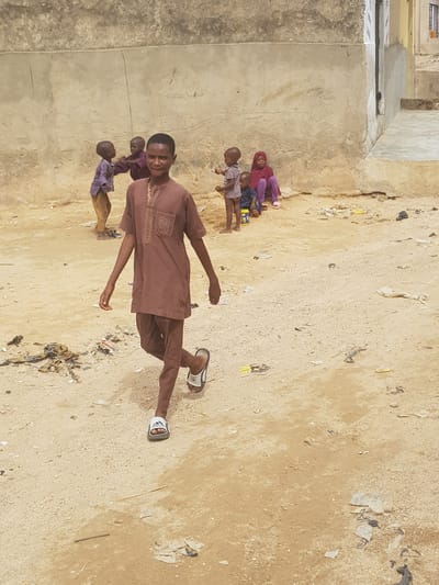 A brown-skinned man wearing a brown shirt and pants, with sandals, is standing in a dirt yard with trash scattered about. Three children stand behind him, near a cement wall. Two of the children are wearing purple and one is wearing pink. The location appears to be Dutsinma A, Nigeria.