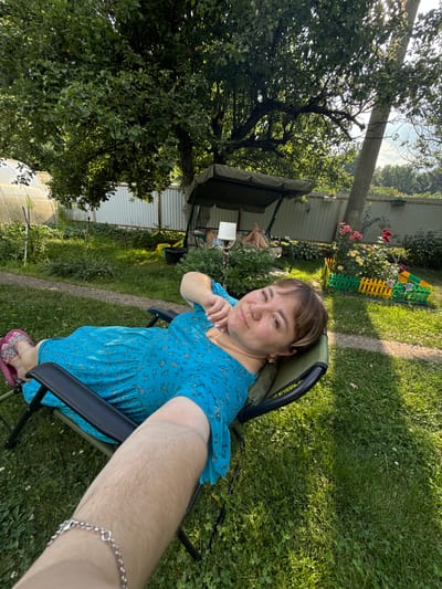 A woman is sitting in a lawn chair in a yard. She is wearing a blue floral dress and pink flip-flops. Her right arm is resting on the armrest of the chair, and she is looking at the camera. The background shows a fence, a swing set, a rose bush, and a greenhouse. The sun is shining, indicating it is daytime. The scene appears to be a backyard in a rural setting. There is no visible text in the image.